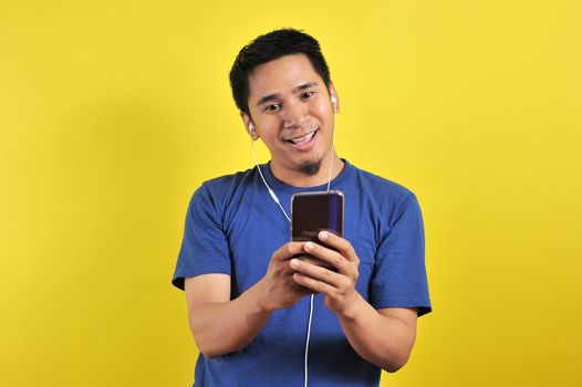 Asian man in casual blue t-shirt wearing headset listening to music from smartphone, isolated on yellow background.