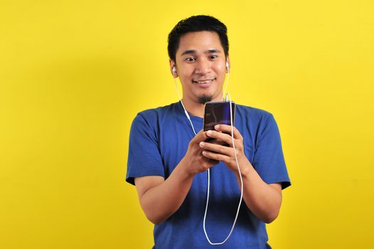 Asian man in casual blue t-shirt wearing headset listening to music from smartphone, isolated on yellow background.