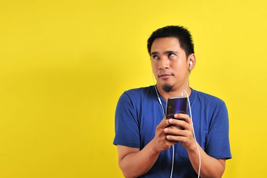 Asian man in casual blue t-shirt wearing headset listening to music from smartphone looking at the copy space, on yellow background.