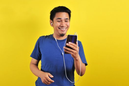 Asian man in casual blue t-shirt happy wearing headset listening to music from smartphone, isolated on yellow background.