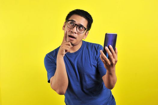 Asian man holding smartphone doing thinking gesture, isolated on yellow background
