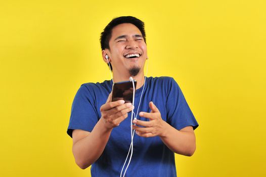 Portrait of excited Asian man laugh happily listening of music from smartphone, isolated on yellow background