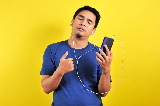 Portrait of Asian man listening of music from smartphone with closed eyes, isolated on yellow background