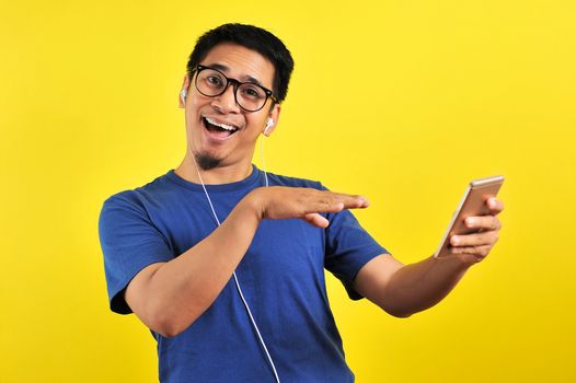 Portrait of excited Asian man laugh happily listening of music from smartphone, isolated on yellow background