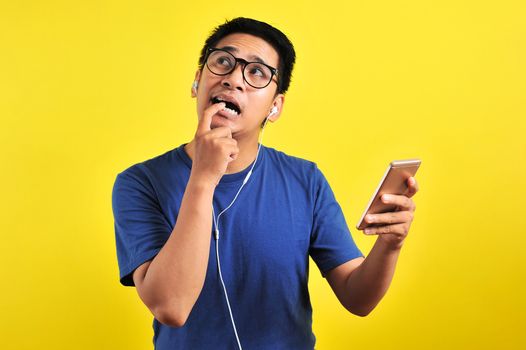 Portrait of confused Young Asian man bitting his finger and listening live music from smartphone with headset, isolated on yellow background