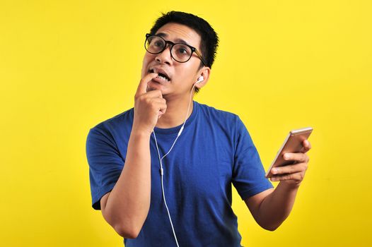 Portrait of confused Young Asian man bitting his finger and listening live music from smartphone with headset, isolated on yellow background
