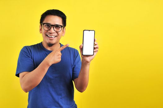 Happy Asian man showing a phone screen and pointing, isolated on yellow background
