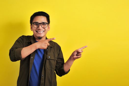 Friendly-looking lively pleasant Asian male with casual t-shirt smiling delighted look at copy space joyful. Self-assured pointing, on yellow background