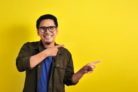 Friendly-looking lively pleasant Asian male with casual t-shirt smiling delighted look at copy space joyful. Self-assured pointing, on yellow background