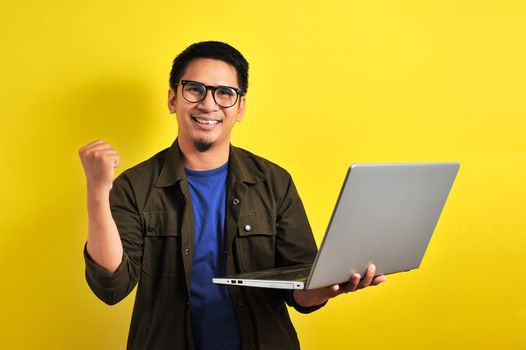 Asian man holding laptop with winning gesture. Asian bussinesman winning gift or lottery, on yellow background