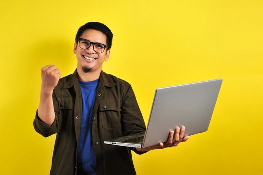 Asian man holding laptop with winning gesture. Asian bussinesman winning gift or lottery, on yellow background