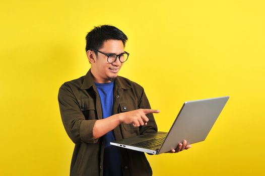 Smiling pleased handsome asian young male entrepreneur, pointing laptop display, isolated on yellow