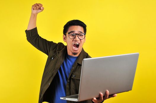 Asian online freelancer very angry in front of his laptop, get bad report from customer, isolated on yellow