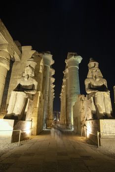 Large statues of Ramses II with columns in hypostyle hall at ancient egyptian Luxor Temple lit up during night