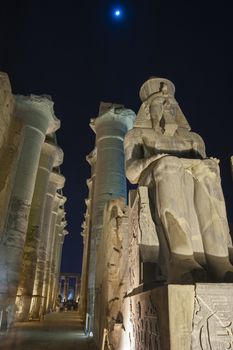 Large statue of Ramses II with columns in hypostyle hall at ancient egyptian Luxor Temple lit up during night