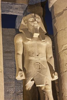 Large statue of Ramses II with columns in hypostyle hall at ancient egyptian Luxor Temple lit up during night
