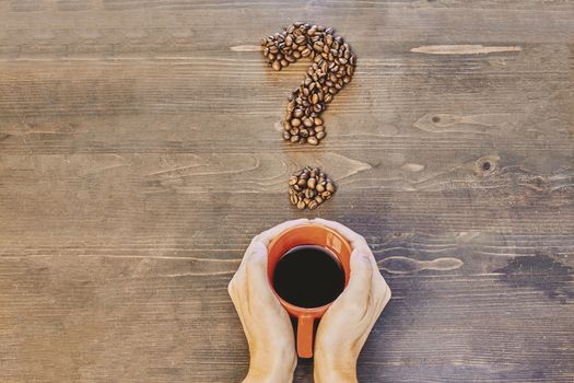 Hands holding a red coffee cup on a wooden table and with a question mark made of coffee beans