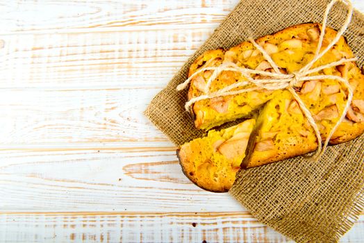 Fresh bakery. Top view of baked pie with apples on sackcloth on a white wooden background. Rustic style.