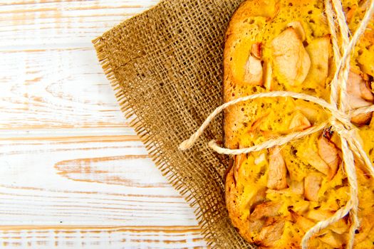 Fresh bakery. Top view of baked pie with apples on sackcloth on a white wooden background. Rustic style.