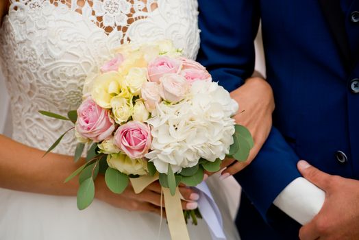 The bride and groom stand with their hands together. The bride's bouquet.