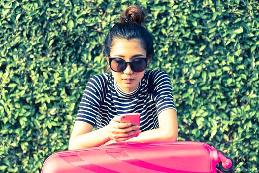 Beautiful traveler asian woman sitting on the street with big pink bag for playing and chatting a mobile phone in the evening with tree background and sunset time.