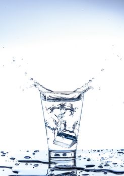 Ice dropping and water splashing in the glass with reflection on the mirror table and isolated on white background.