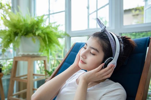 Beautiful Asian women wear headphones and use a notebook computer to work and listen to music in the garden at home on a relaxing day.