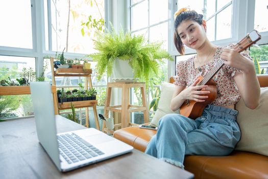 Asian women use their notebook computers to study and practice playing ukulele on the internet at home.
