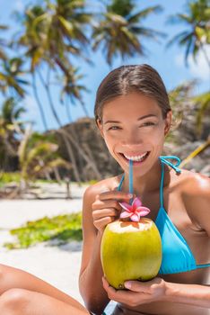 Beautiful Asian bikini woman drinking fresh coconut water directly from fruit with straw on beach. Multiracial girl smiling at camera portrait with perfect teeth sipping on hydration juice food.