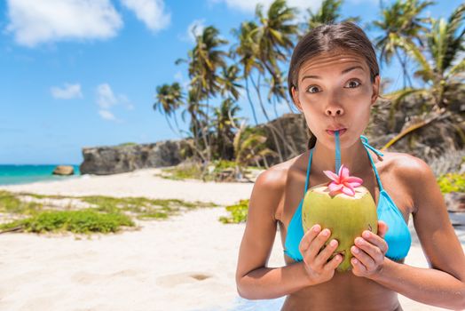 Funny girl having fun drinking coconut water on beach. Cute Asian woman doing goofy face sipping with straw on natural fruit for healthy hydration drink during tropical travel destination holiday.