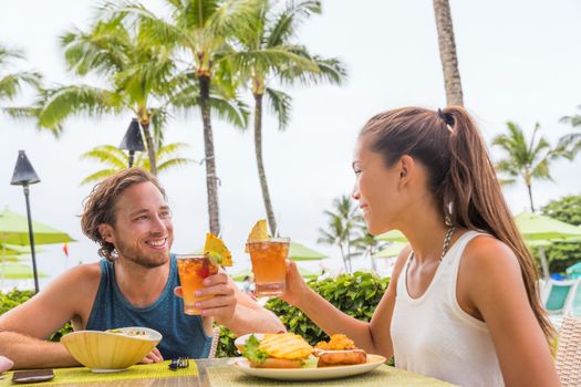 Couple eating at hotel restaurant on Hawaii travel vacation beach drinking hawaiian drink mai tai. Happy people toasting cheers with cocktails. Summer holidays at resort.