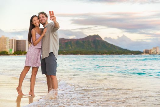 Couple walking on beach at sunset taking selfie picture on mobile phone relaxing together on Waikiki beach, Honolulu, Hawaii travel vacation. Romantic holiday destination for honeymoon.