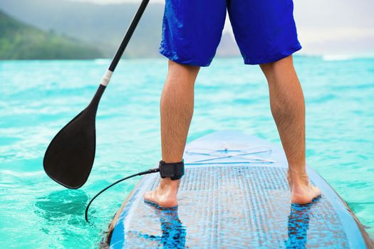 Paddle board man doing stand-up paddleboard on ocean. Athlete paddleboarding on SUP surf board on Hawaii beach travel. Closeup of legs standing on board.