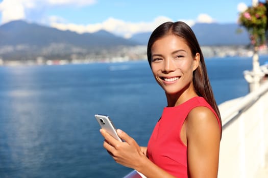 Happy asian woman using phone app in vancouver city harbour on summer day. Beautiful landscape of canadian nature river and mountains. Businesswoman on smartphone lifestyle.