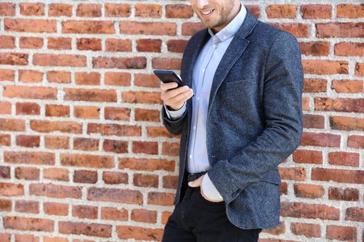 Young casual business man using smartphone in urban city background relaxing on brick wall texting sms on phone app living a modern lifestyle. Closeup of unrecognizable person.