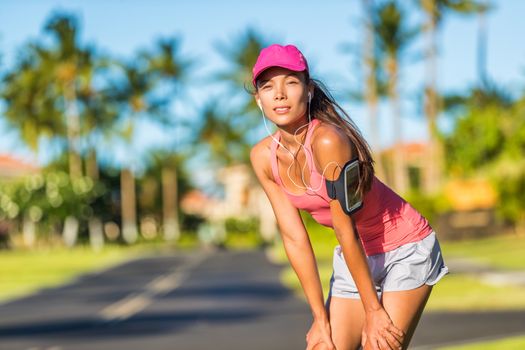 Tired runner woman ready for running wearing sports cap and sport armband with earphones listening to mobile music. Active fit Asian girl resting taking a jogging break on outdoor summer street.