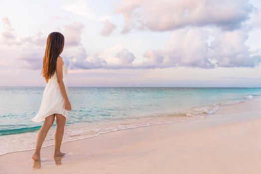 Peaceful vacation paradise woman walking on sunset beach with pastel colors sky and ocean for tranquility and serenity . Girl in white dress relaxing on luxury tropical summer getaway