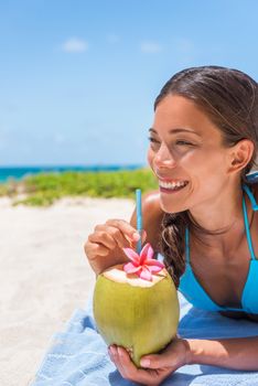 Happy Asian girl drinking fresh coconut water on beach vacation. Tropical travel destination, Sun tanning in summer holidays. Tourist woman relaxing enjoying drink.