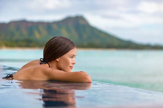 Luxury vacation travel woman relaxing in infinity swimming pool on summer beach resort. Asian girl tourist on wellness spa relaxation watching sunset outside in nature landscape background.