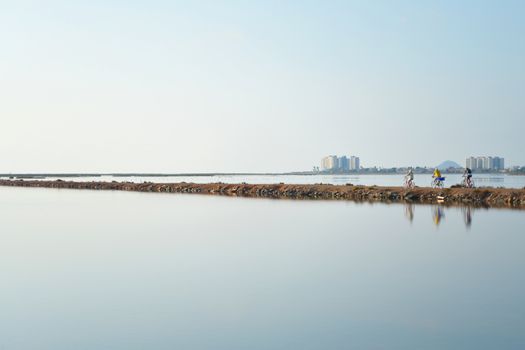 Gentle evening colors surround 3 pedal bike cyclists as they cycle along a thin strip of land between lagoons. Reflections shimmer in still water.