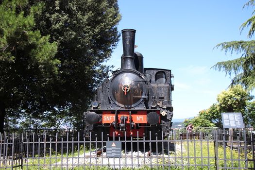 Locomotiva a vapore SNFT - N.1 steam locomotive Prisoner monument on Cidneo Hill of Castle in green park, historical city centre, green trees blue sky, Lombardy