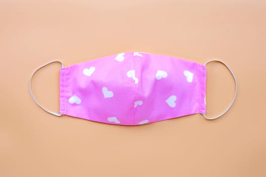 Handmade pink cloth mask with white heart shape on orange background. Top view