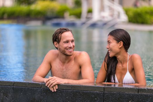 Pool couple relaxing on luxury resort travel holiday talking together on summer vacation. Asian woman, caucasian man in love.