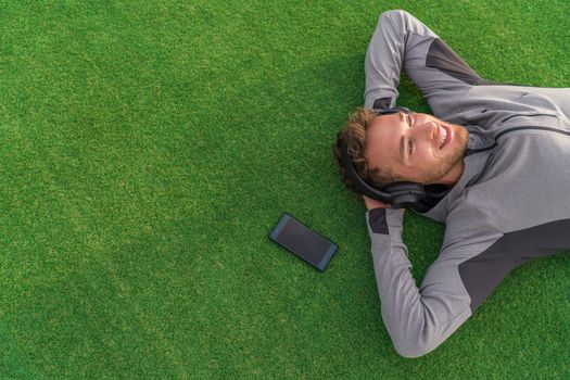 Happy man listening to music with headphones and phone relaxing on green grass lying down with arms behind head enjoying summer day in park. Young adult using mindfulness smartphone app concept.