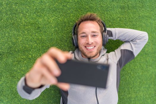 Happy man watching phone video app listening with headphones and smartphone relaxing outside on summer green grass.