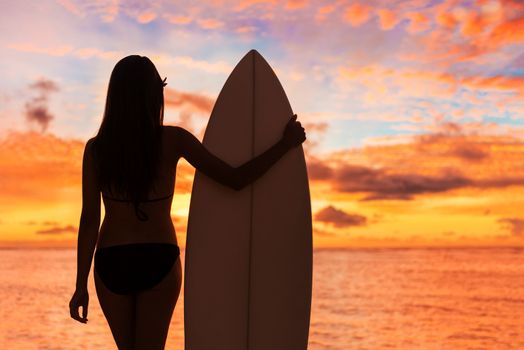 Beach sunset sexy surfer woman surfing lifestyle relaxing holding surfboard looking at ocean waves for surf. Active healthy living silhouette of sports athlete standing in colorful sky.