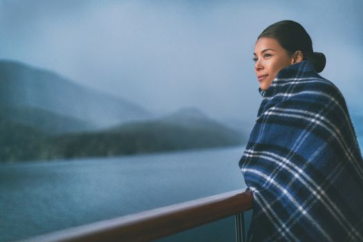 Cruise ship passenger on Alaska travel vacation enjoying scenery at dusk on suite balcony deck with wool throw in cold weather. Asian tourist woman relaxing on summer holiday cruising adventure.