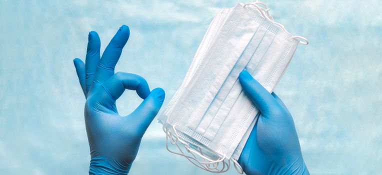 Close-up of doctor hands in blue medical gloves holding surgical medical face mask, showing okay sign. Banner panorama medical concept of protection against coronavirus, infection, bacteria and virus.