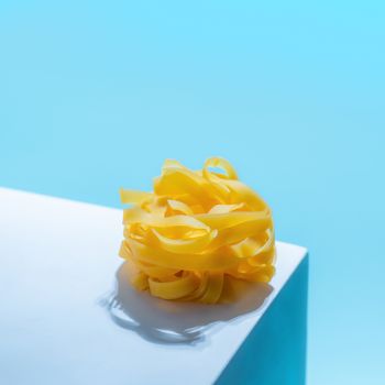 Pasta art. Tagliatelle on blue pastel pedestal over blue background in hard light. Creative shot of pasta noodle. Creative set design for menu of italian cuisine with copy space