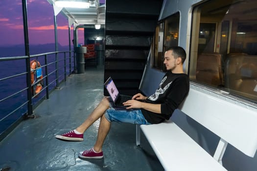 A young man works on a laptop on the deck of a ship. Remote work. Always in touch